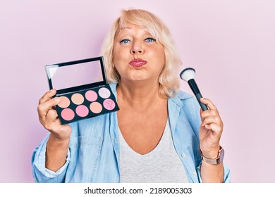 Middle Age Blonde Woman Holding Makeup Brush And Blush Puffing Cheeks With Funny Face. Mouth Inflated With Air, Catching Air. 