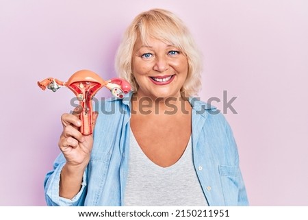 Middle age blonde woman holding anatomical model of female genital organ looking positive and happy standing and smiling with a confident smile showing teeth 