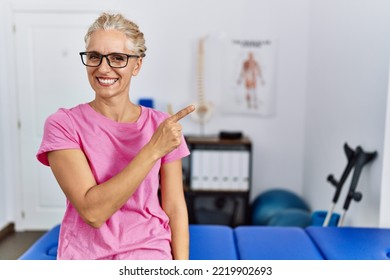 Middle Age Blonde Woman At Pain Recovery Clinic Cheerful With A Smile Of Face Pointing With Hand And Finger Up To The Side With Happy And Natural Expression On Face 