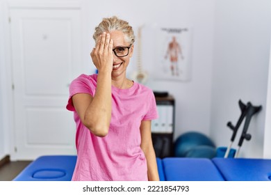 Middle Age Blonde Woman At Pain Recovery Clinic Covering One Eye With Hand, Confident Smile On Face And Surprise Emotion. 