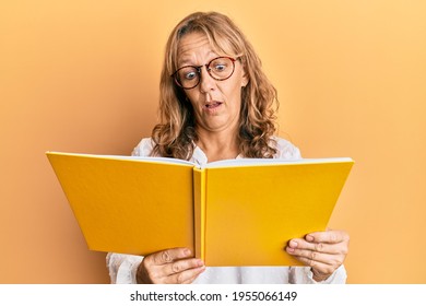 Middle Age Blonde Woman Reading A Book Wearing Glasses In Shock Face, Looking Skeptical And Sarcastic, Surprised With Open Mouth 