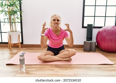 Middle Age Blonde Woman Sitting On Yoga Mat Shouting Frustrated With Rage, Hands Trying To Strangle, Yelling Mad 