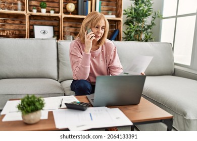 Middle age blonde woman teleworking using laptop and talking on the smartphone at home. - Powered by Shutterstock