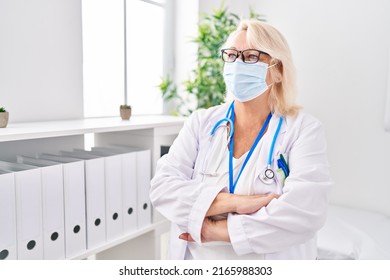 Middle age blonde woman wearing doctor uniform and medical mask standing with arms crossed gesture at clinic