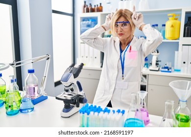 Middle Age Blonde Woman Working At Scientist Laboratory Doing Funny Gesture With Finger Over Head As Bull Horns 