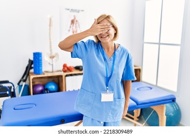 Middle Age Blonde Woman Working At Pain Recovery Clinic Smiling And Laughing With Hand On Face Covering Eyes For Surprise. Blind Concept. 