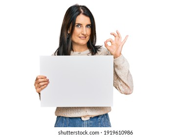 Middle age brunette woman holding blank empty banner doing ok sign with fingers, smiling friendly gesturing excellent symbol 