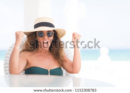 Middle age brunette woman sitting at the table by the beach annoyed and frustrated shouting with anger, crazy and yelling with raised hand, anger concept