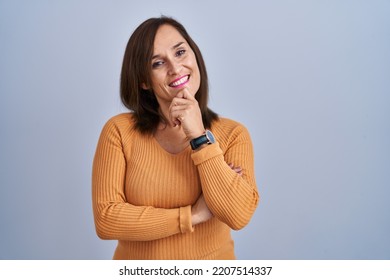 Middle Age Brunette Woman Standing Wearing Orange Sweater With Hand On Chin Thinking About Question, Pensive Expression. Smiling And Thoughtful Face. Doubt Concept. 