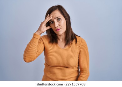 Middle age brunette woman standing wearing orange sweater worried and stressed about a problem with hand on forehead, nervous and anxious for crisis  - Powered by Shutterstock