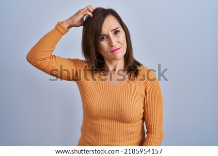 Middle age brunette woman standing wearing orange sweater confuse and wondering about question. uncertain with doubt, thinking with hand on head. pensive concept. 