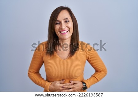 Middle age brunette woman standing wearing orange sweater smiling and laughing hard out loud because funny crazy joke with hands on body. 