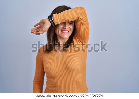 Middle age brunette woman standing wearing orange sweater covering eyes with arm smiling cheerful and funny. blind concept. 