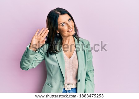 Middle age brunette woman wearing casual clothes waiving saying hello happy and smiling, friendly welcome gesture 