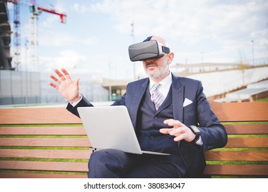 Middle age caucasian business man sitting on a bench with laptop on his knee using smart watch and 3D viewer - futuristic, multitasking, technology concept - Powered by Shutterstock