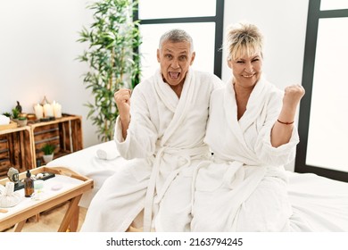 Middle age caucasian couple wearing bathrobe at wellness spa screaming proud, celebrating victory and success very excited with raised arms 