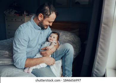 Middle Age Caucasian Father Holding Rocking Newborn Baby On Arms. Tired Man Parent Sitting On Bed At Home With Child In Hands. Authentic Lifestyle Family Moment. Single Dad Home Life. 