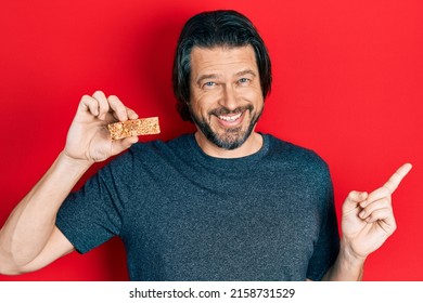 Middle Age Caucasian Man Eating Protein Bar As Healthy Energy Snack Smiling Happy Pointing With Hand And Finger To The Side 