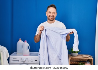 Middle Age Caucasian Man Holding Shirt With Sweat Winking Looking At The Camera With Sexy Expression, Cheerful And Happy Face. 