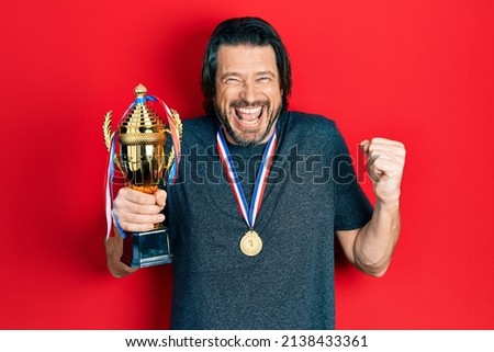 Middle age caucasian man holding champion trophy wearing medals screaming proud, celebrating victory and success very excited with raised arm 