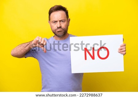 Middle age caucasian man isolated on yellow background holding a placard with text NO and doing bad signal