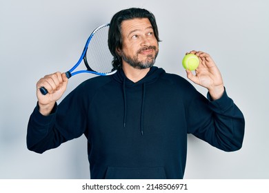 Middle Age Caucasian Man Playing Tennis Holding Racket And Ball Smiling Looking To The Side And Staring Away Thinking. 