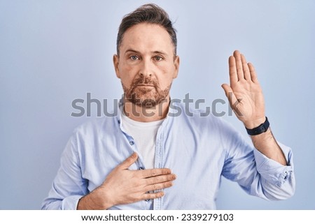Middle age caucasian man standing over blue background swearing with hand on chest and open palm, making a loyalty promise oath 