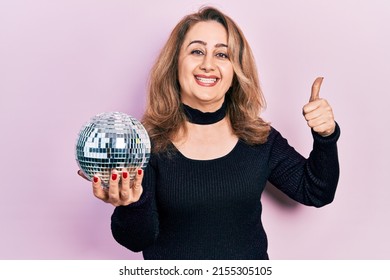 Middle age caucasian woman holding shiny disco ball smiling happy and positive, thumb up doing excellent and approval sign 