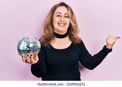 Middle age caucasian woman holding shiny disco ball pointing thumb up to the side smiling happy with open mouth 