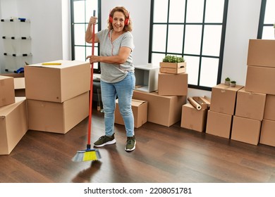 Middle Age Caucasian Woman Listening To Music Cleaning Floor Using Broom At New Home.