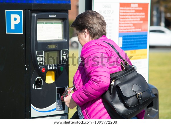 Middle age caucasian woman\
paying for paying at a parking meter in a car park outside on a\
sunny day