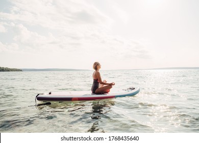Middle age Caucasian woman practising yoga on paddle sup surfboard at sunset. Female stretching doing workout on lake water. Modern individual hipster outdoor summer sport activity.