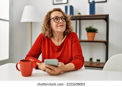 Middle age caucasian woman using smartphone and drinking coffee sitting on the sofa at home. - Shutterstock ID 2222638001