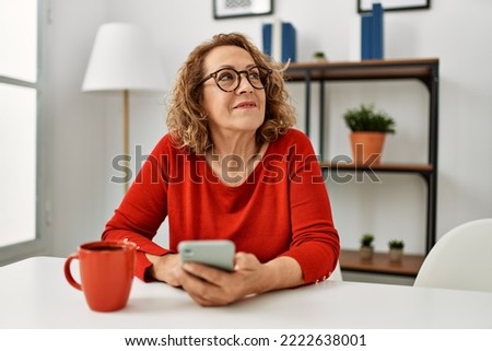 Middle age caucasian woman using smartphone and drinking coffee sitting on the sofa at home.