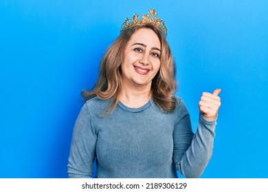 Middle Age Caucasian Woman Wearing Queen Crown Smiling With Happy Face Looking And Pointing To The Side With Thumb Up. 