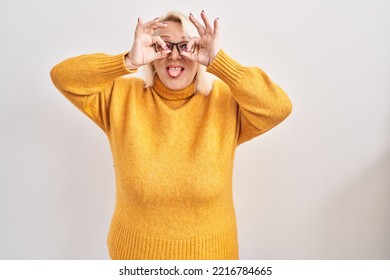 Middle Age Caucasian Woman Wearing Glasses Standing Over Background Doing Ok Gesture Like Binoculars Sticking Tongue Out, Eyes Looking Through Fingers. Crazy Expression. 