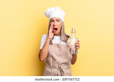 Middle Age Chef Woman Feeling Happy, Excited And Positive, Giving A Big Shout Out With Hands Next To Mouth, Calling Out With A Water Bottle