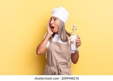 Middle Age Chef Woman Feeling Happy, Excited And Surprised, Looking To The Side With Both Hands On Face With A Water Bottle