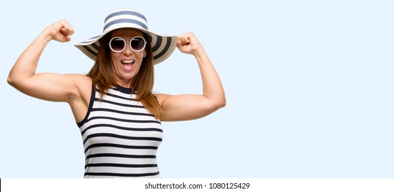 Middle age cool woman wearing summer hat and sunglasses happy and excited celebrating victory expressing big success, power, energy and positive emotions. Joyful isolated blue background