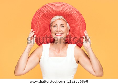 Middle age cool woman wearing red summer hat looking at camera and smiling, posing on yellow studio background.