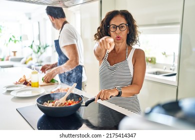 Middle Age Couple Cooking Mediterranean Food At Home Pointing With Finger To The Camera And To You, Confident Gesture Looking Serious 
