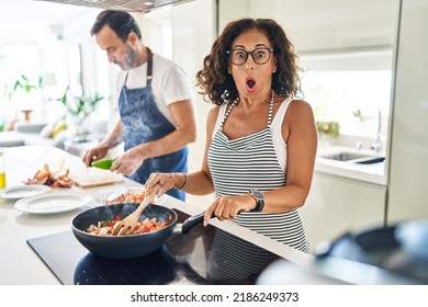 Middle Age Couple Cooking Mediterranean Food At Home Scared And Amazed With Open Mouth For Surprise, Disbelief Face 