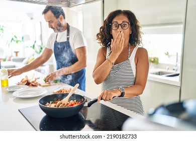 Middle Age Couple Cooking Mediterranean Food At Home Covering Mouth With Hand, Shocked And Afraid For Mistake. Surprised Expression 