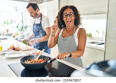 Middle Age Couple Cooking Mediterranean Food At Home Surprised With An Idea Or Question Pointing Finger With Happy Face, Number One 