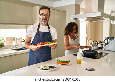 Middle Age Couple Cooking Mediterranean Food At Home Relaxed With Serious Expression On Face. Simple And Natural Looking At The Camera. 