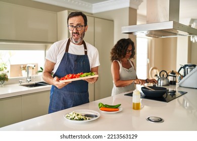 Middle Age Couple Cooking Mediterranean Food At Home Clueless And Confused Expression. Doubt Concept. 