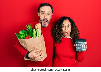 Middle Age Couple Of Hispanic Woman And Man Holding Groceries Bag And Calculator Afraid And Shocked With Surprise And Amazed Expression, Fear And Excited Face. 