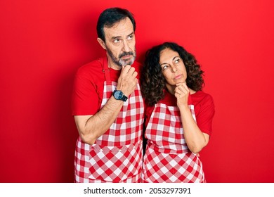Middle age couple of hispanic woman and man wearing cook apron with hand on chin thinking about question, pensive expression. smiling with thoughtful face. doubt concept. 