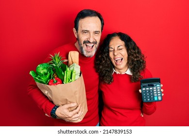 Middle Age Couple Of Hispanic Woman And Man Holding Groceries Bag And Calculator Smiling And Laughing Hard Out Loud Because Funny Crazy Joke. 