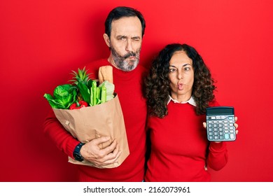 Middle Age Couple Of Hispanic Woman And Man Holding Groceries Bag And Calculator Making Fish Face With Mouth And Squinting Eyes, Crazy And Comical. 